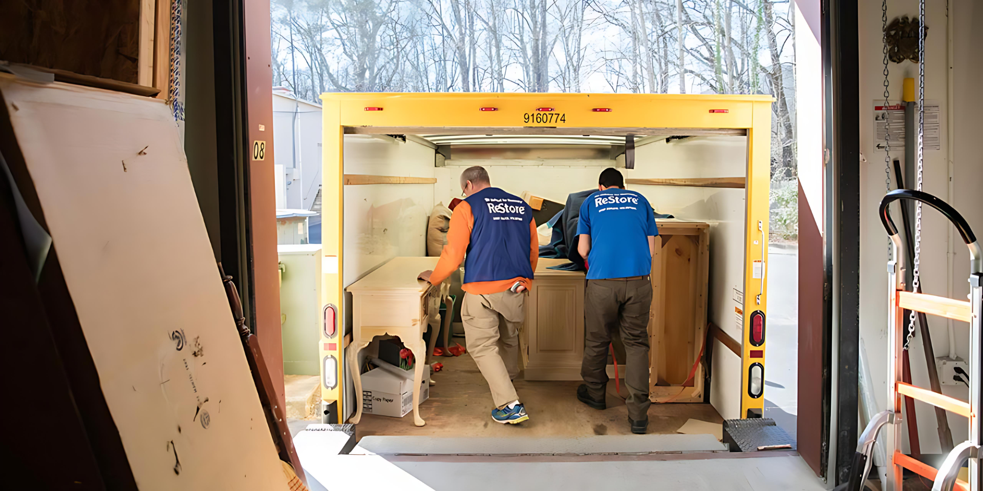 Two men wearing work gloves and blue 'ReStore' branded vests move a vanity, sideboard, and other furniture into/out of a truck on a snowy day