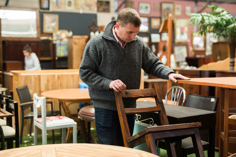 Man browsing a furniture store looks down at a chair, resting one hand on it and another on a table