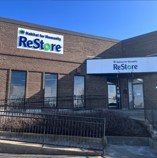 Addison Habitat for Humanity ReStore storefront with blue and green "Habitat for Humanity ReStore" sign, a brick exterior, concrete stairs and wheelchair ramp, and slim glass windows under a clear blue sky.