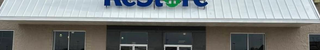 Storefront of the Naperville ReStore on a clear day. It has a white roof, two glass double doors, a visible blue and green "Habitat for Humanity ReStore" sign, and four glass windows with a beige concrete exterior.