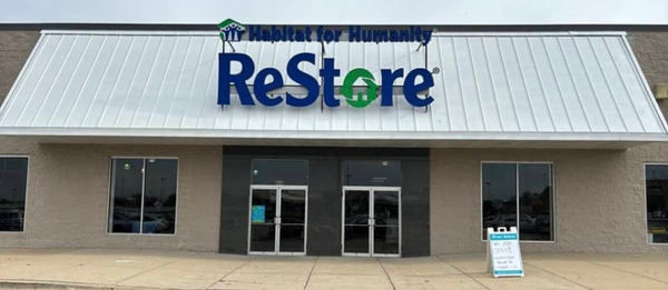 Storefront of the Naperville ReStore on a clear day. It has a white roof, two glass double doors, a visible blue and green "Habitat for Humanity ReStore" sign, and four glass windows with a beige concrete exterior.