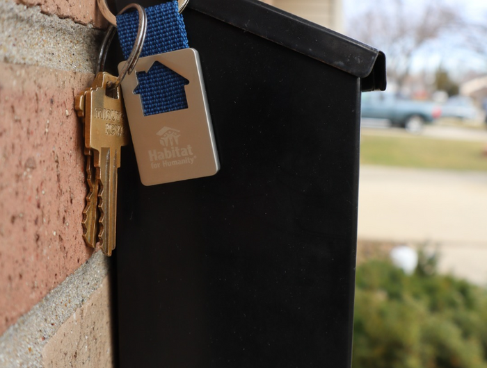 Black wall-mounted mailbox with keys and Habitat for Humanity keychain, with house-shaped cutout, dangling from it.