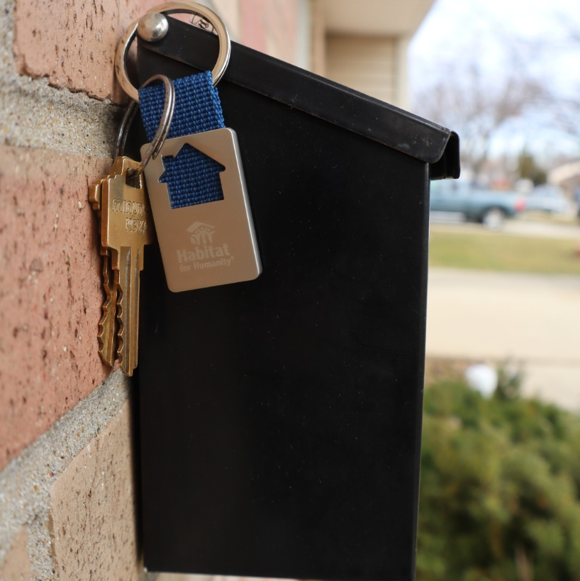 Black wall-mounted mailbox with keys and Habitat for Humanity keychain, with house-shaped cutout, dangling from it.